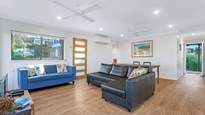 A spacious living room with a blue sofa, a dark leather sofa, and a wooden dining table. There are ceiling fans, a large window with a view of greenery, and a door leading outside. - Banksia House allows you to surround yourself with nature in this serene piece of paradise (Sanctuary Point)
