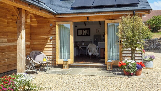 A wooden cabin with a covered patio, solar panels on the roof, and a small garden area with potted plants. - 1 bedroom accommodation in Brighstone (Brighstone)