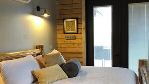 A bed with a white duvet and multiple pillows, a wall-mounted air conditioner, a framed picture, and a wooden door leading to an outdoor area. - Lookout cabin (Kamiah)