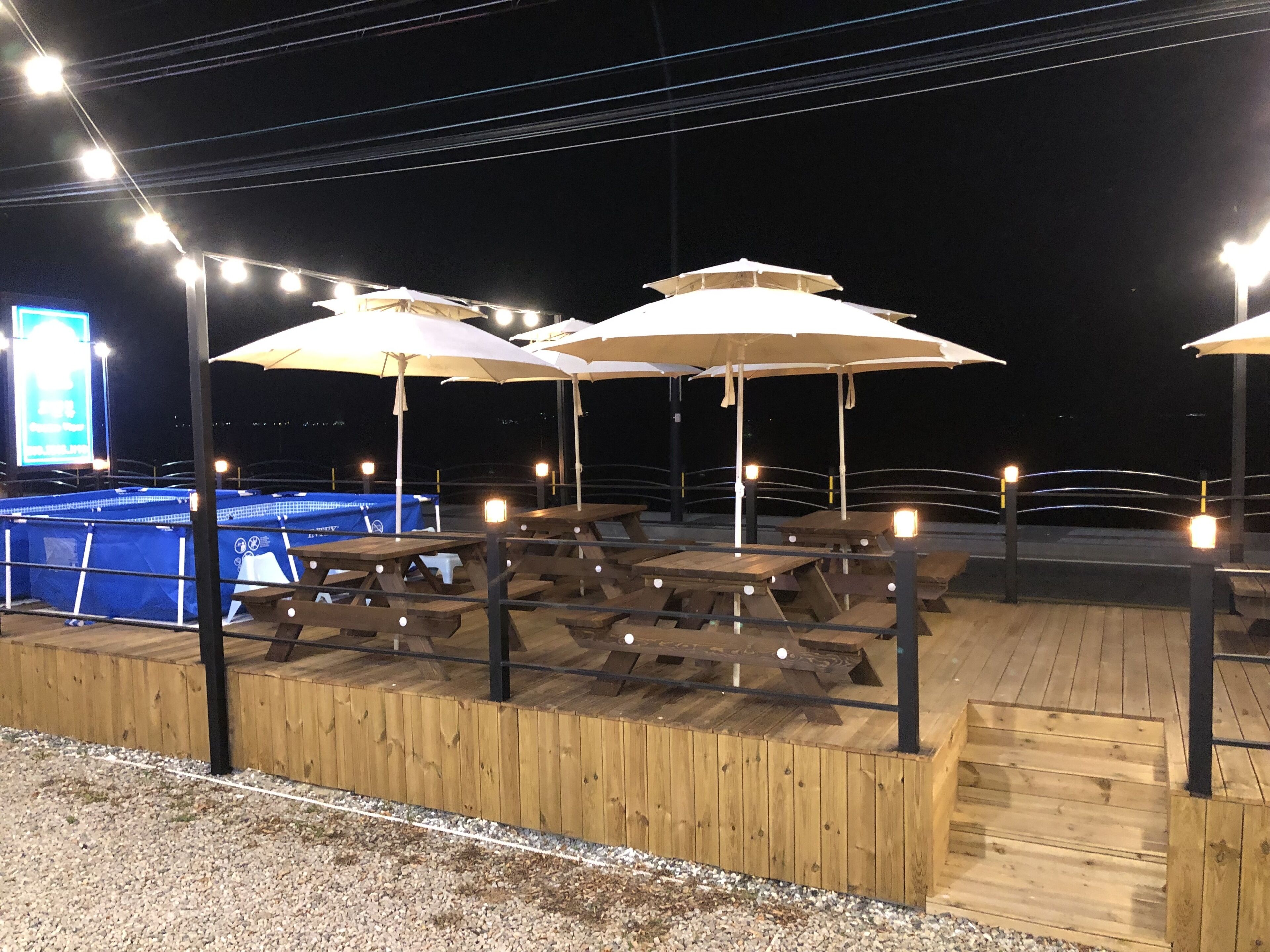 An outdoor seating area with wooden tables and umbrellas, a pool, and a large illuminated sign in the background.