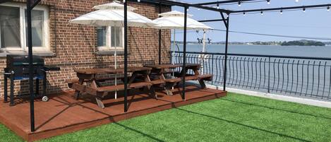 An outdoor patio with picnic tables and umbrellas, a grill, and a view of a body of water.