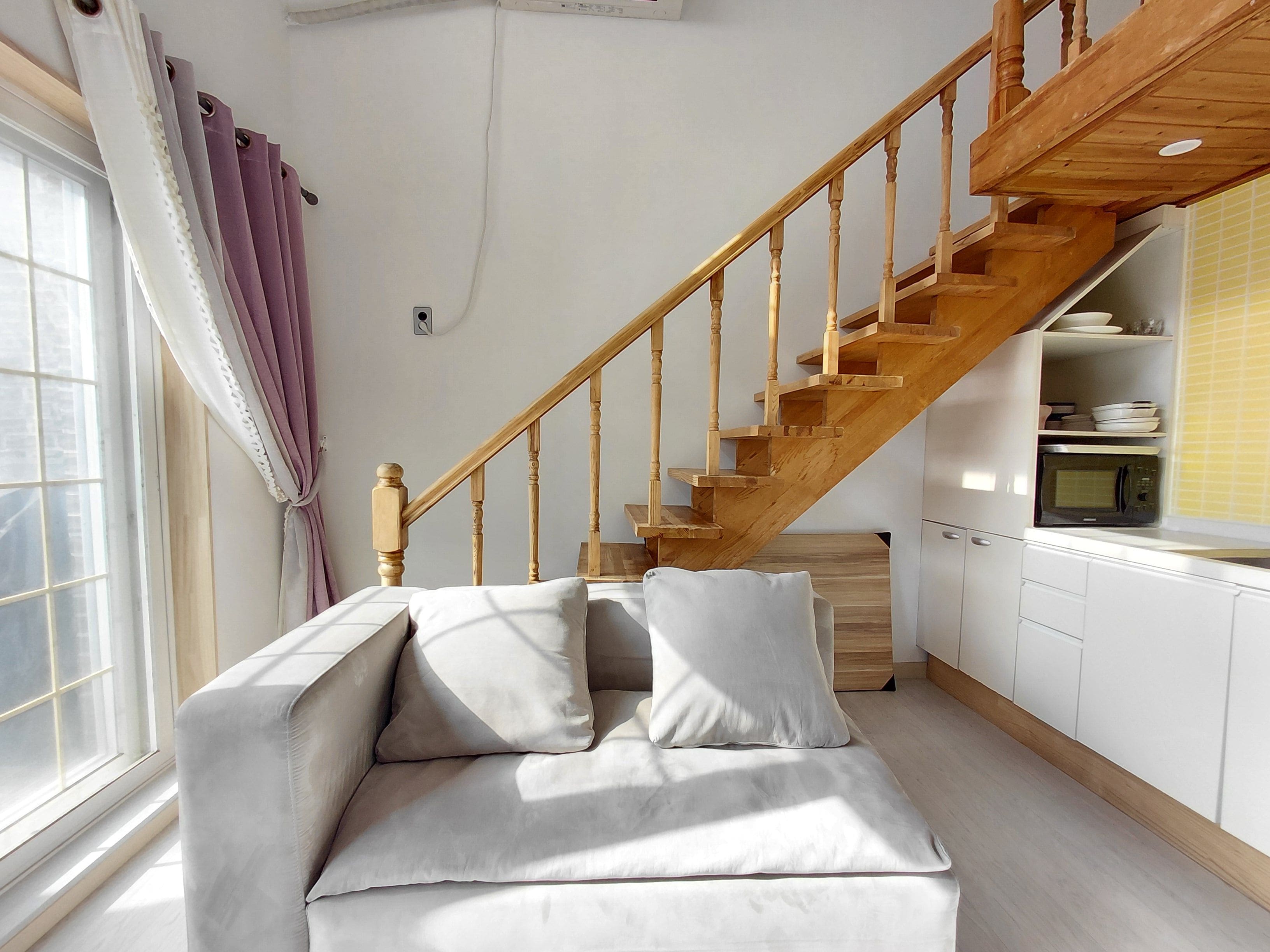 A modern living room with a wooden staircase, a sofa with cushions, and a kitchen area with a microwave.