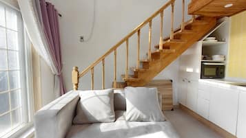 A modern living room with a wooden staircase, a sofa with cushions, and a kitchen area with a microwave.