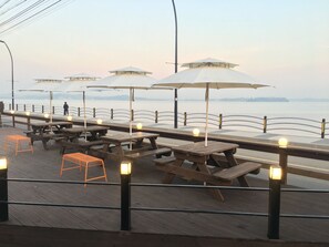 Ein Essbereich am Wasserufer mit Picknicktischen und Sonnenschirmen, mit Blick auf ein Gewässer.