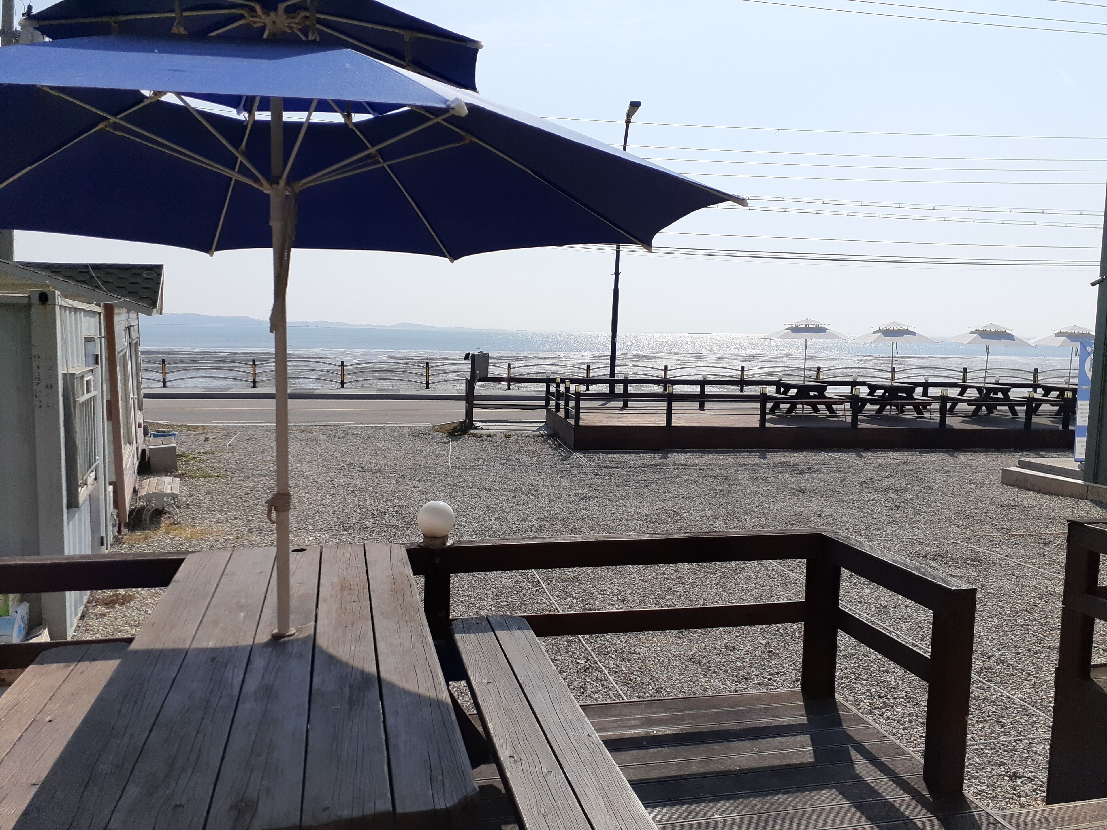 Eine Holzdar Terrasse mit Bänken und einem Sonnenschirm, mit Blick auf einen Kiesstrand mit einem Zaun und Sonnenschirmen in der Ferne.