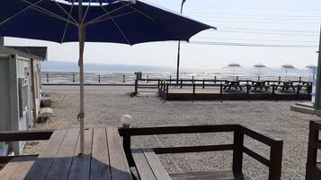 Eine Holzdar Terrasse mit Bänken und einem Sonnenschirm, mit Blick auf einen Kiesstrand mit einem Zaun und Sonnenschirmen in der Ferne.