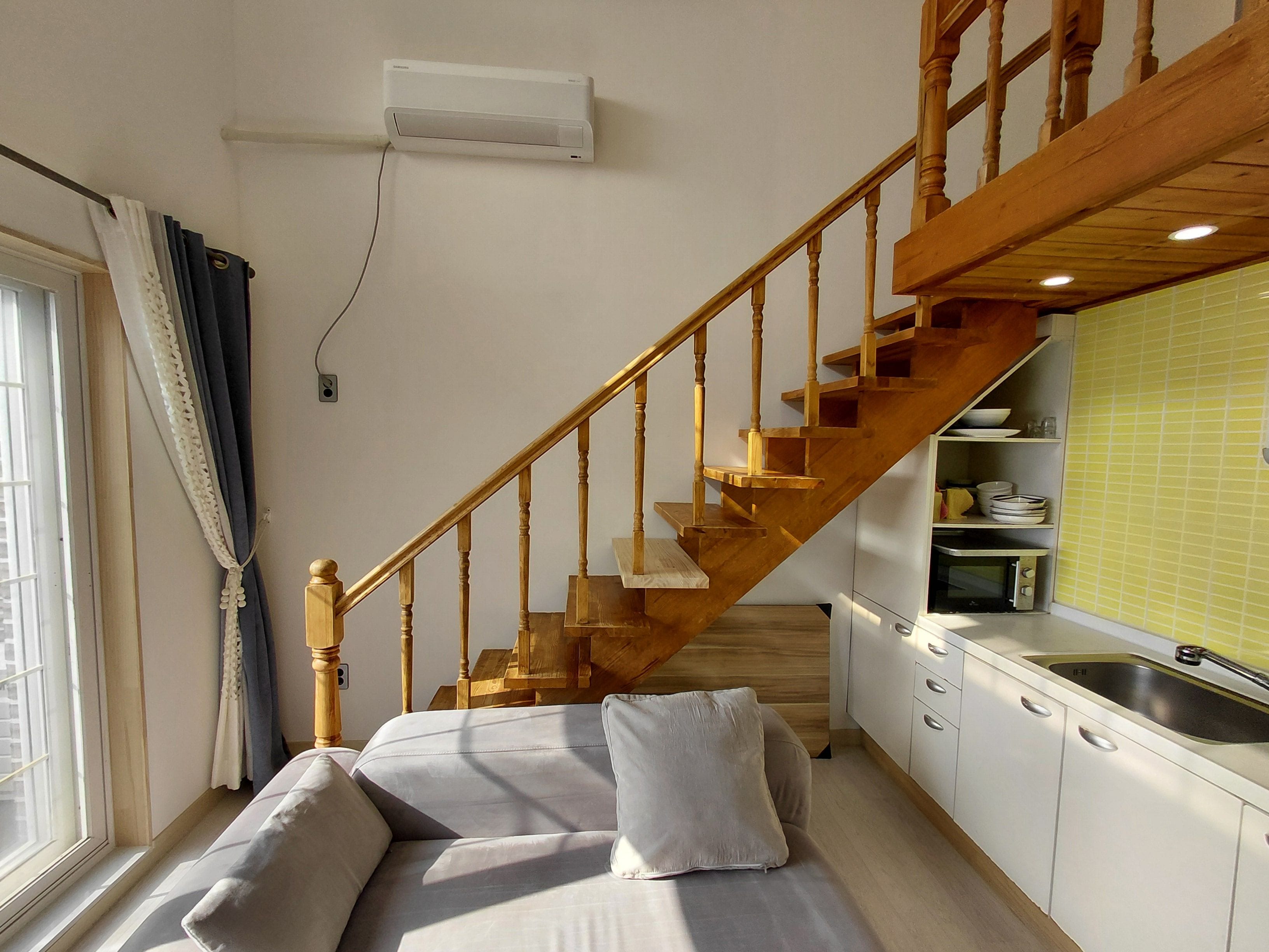 Un espacio habitable pequeño con una escalera de madera, un sofá con cojines, una zona de cocina con un lavabo y una ventana con cortinas.