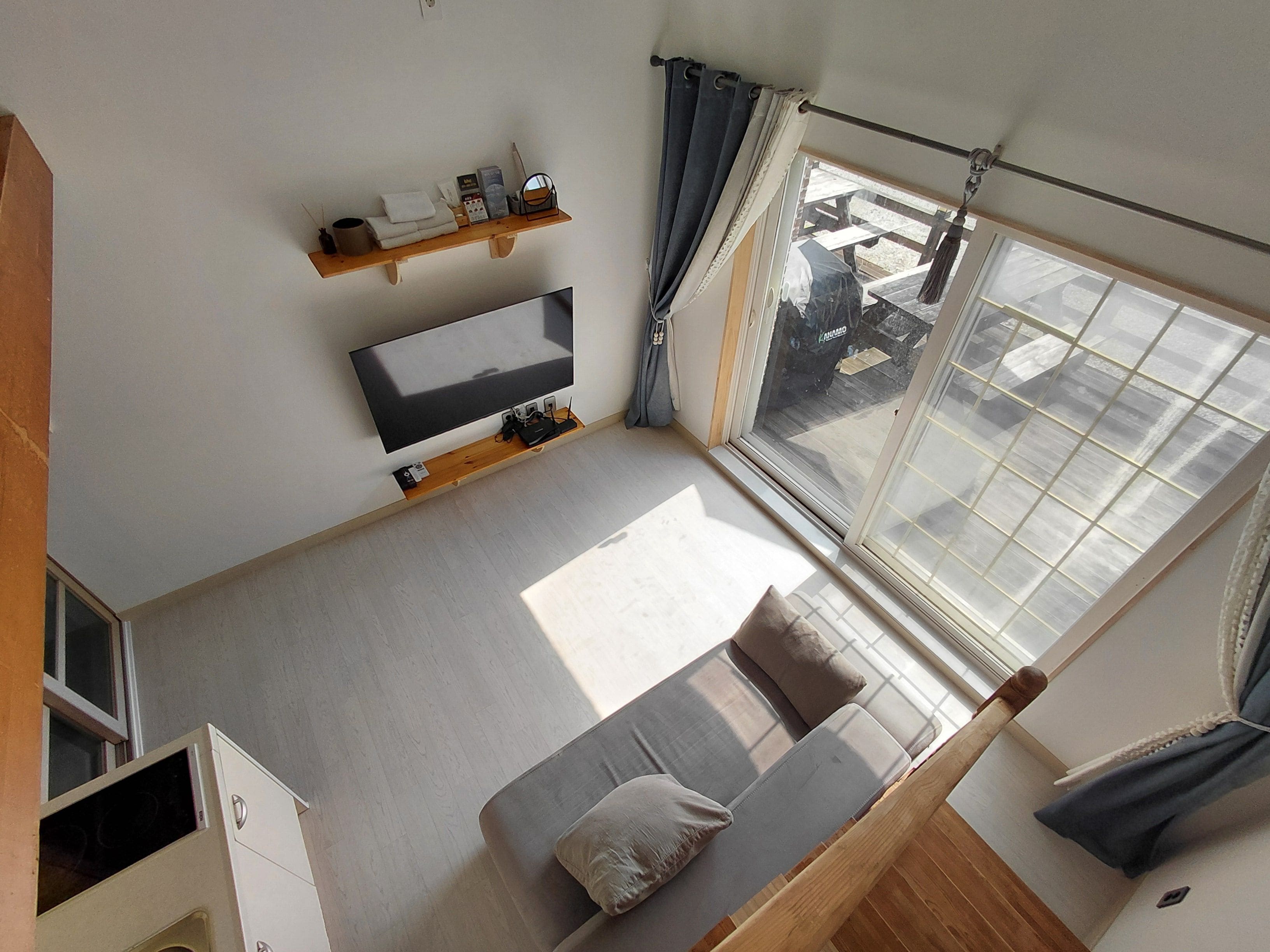 A modern room with a flat-screen TV, a sofa, a wooden shelf with items, and a large window.