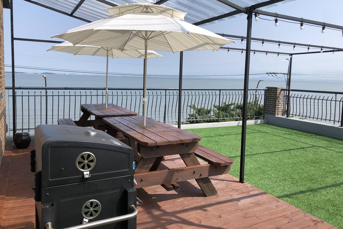 A covered patio with a grill, picnic table, and umbrella, overlooking a body of water.