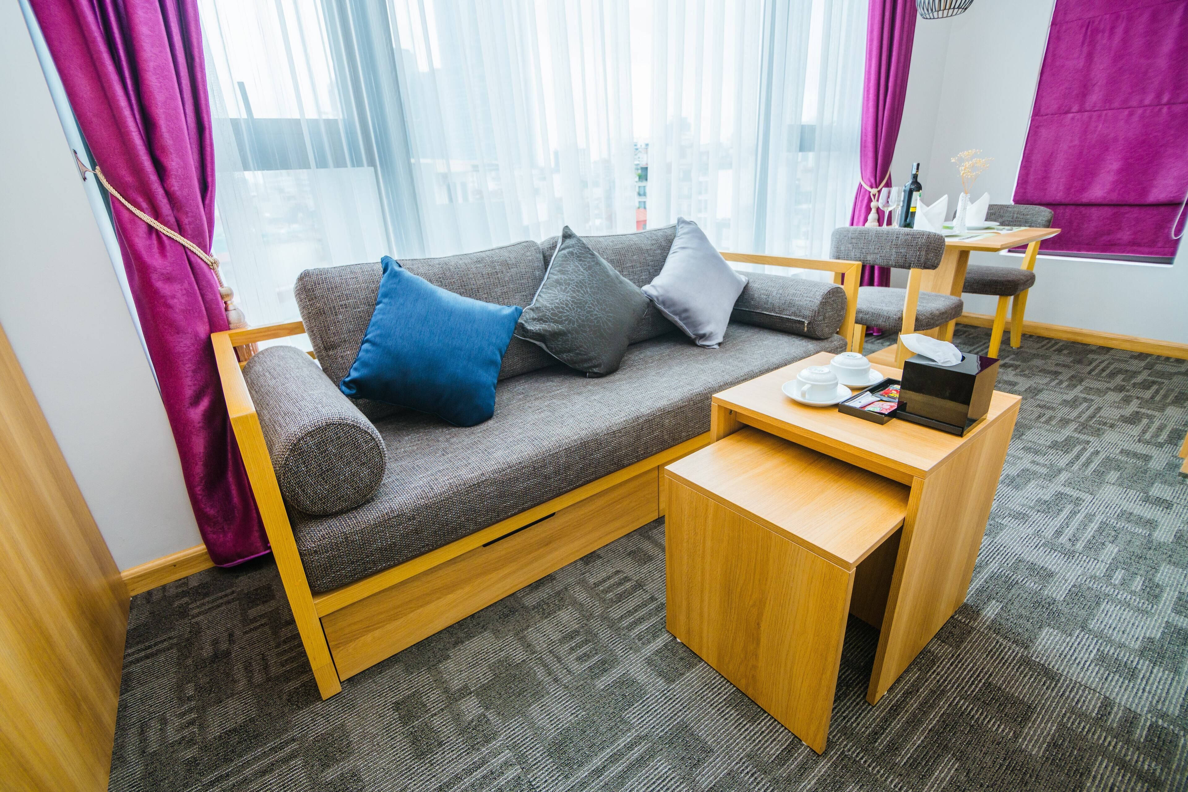 A modern room with a grey sofa, wooden side table, and a view of the city through large windows with purple curtains.