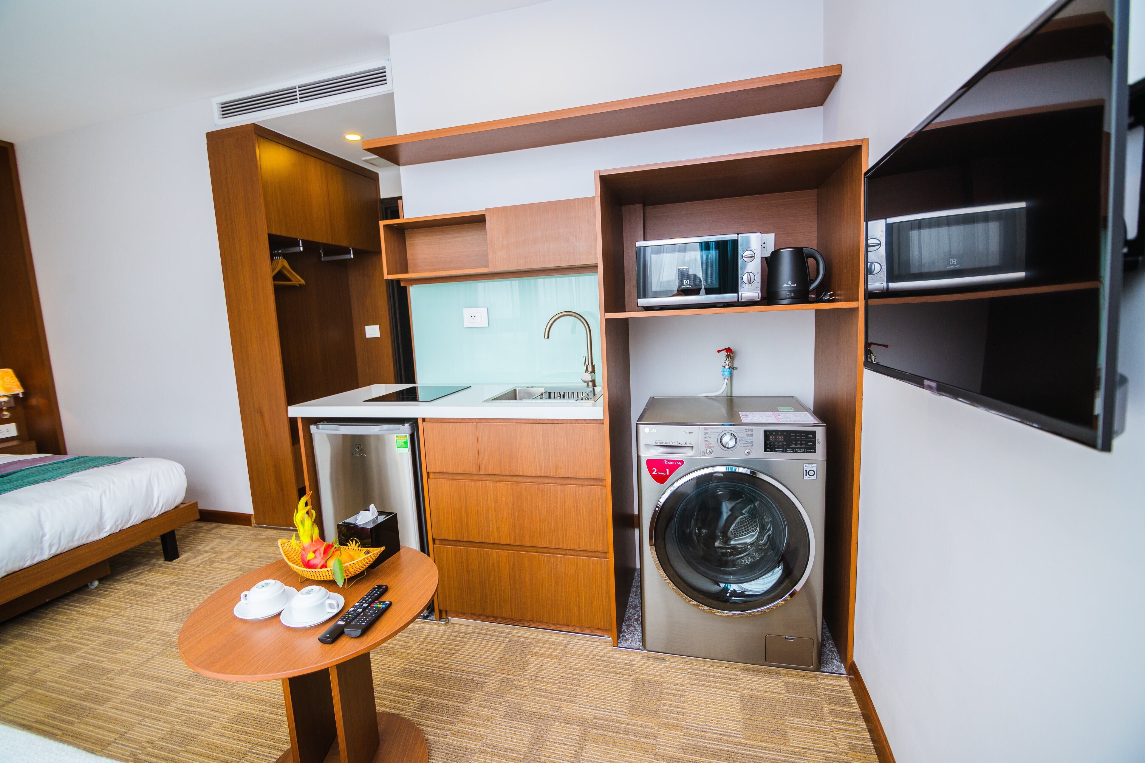 A compact hotel room with a bed, a small table with cups and a remote control, a washing machine, a microwave, and a sink.