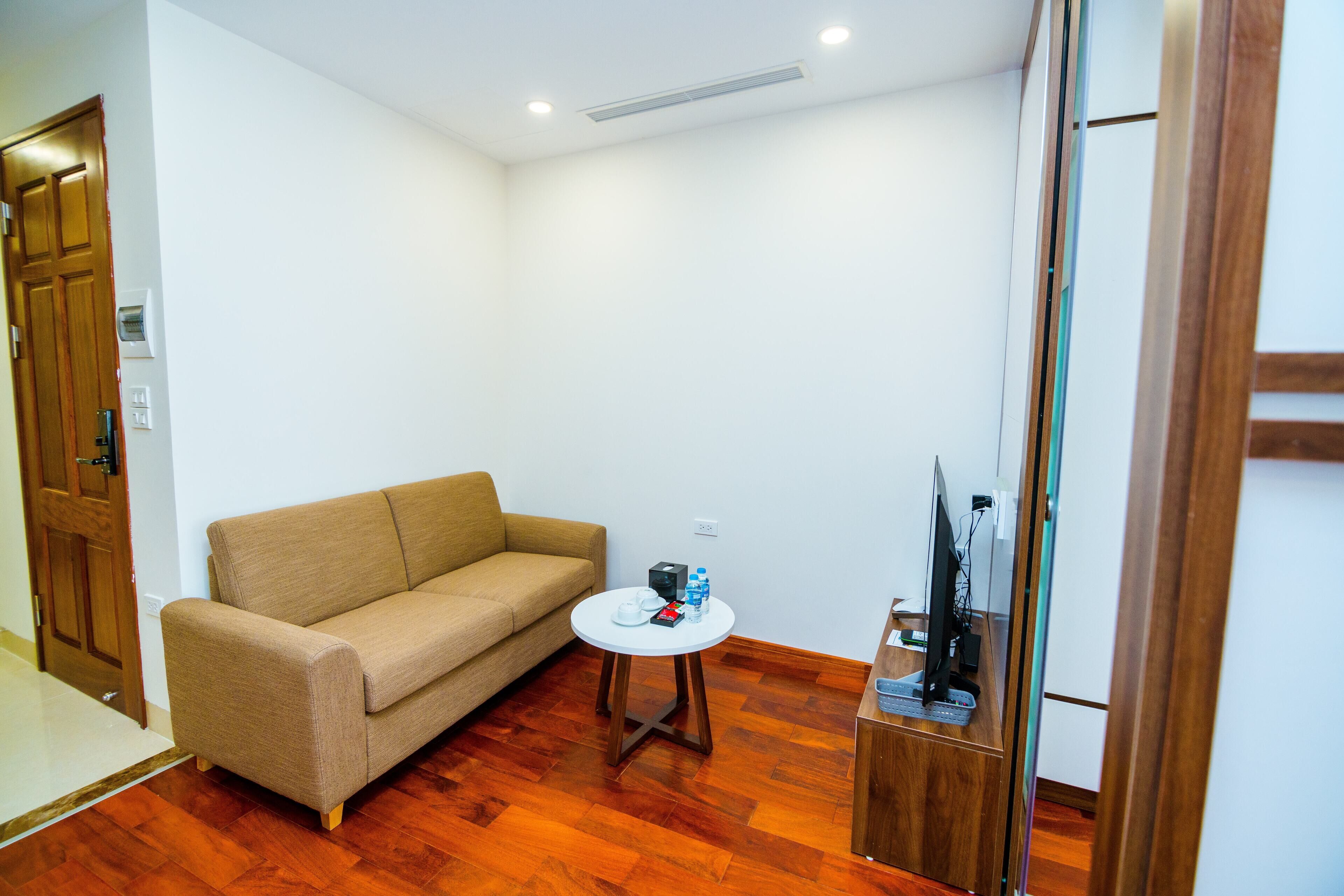 Um quarto pequeno e mobiliado com piso de madeira, um sofá bege, uma mesinha redonda com alguns objetos e uma televisão em um suporte de madeira.