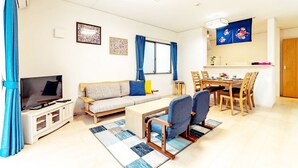 Ein Wohnzimmer mit Sofa, Fernseher, Esstisch, Stühlen und einer Küchenzeile.