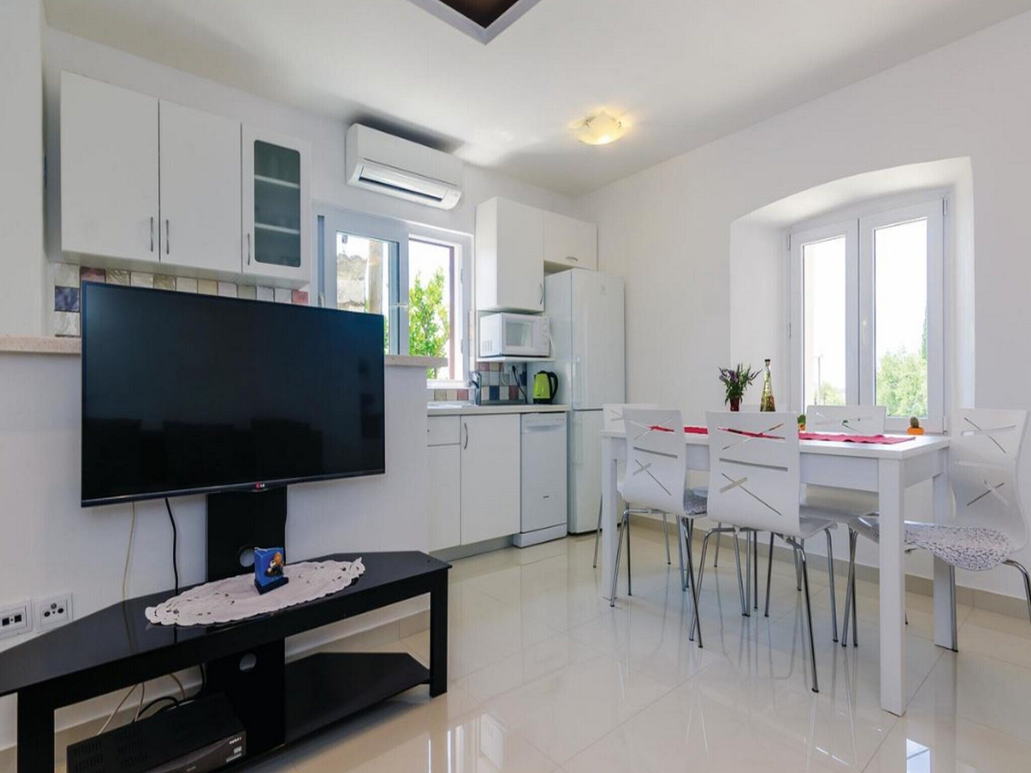 Une cuisine moderne avec des meubles blancs, un téléviseur à écran plat, un coin repas avec des chaises blanches et une fenêtre laissant entrer la lumière naturelle.
