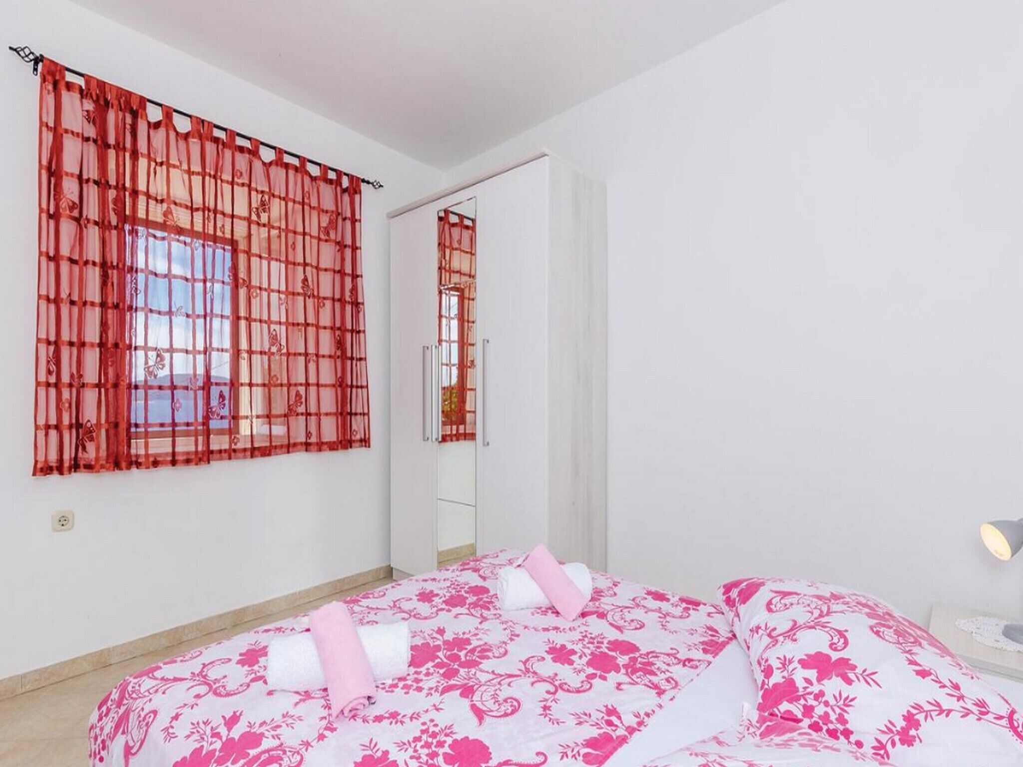 A bedroom with a bed, a window with a red curtain, a mirror, and a bedside table with a lamp.