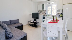 Ein modernes Wohnzimmer mit grauem Sofa, einem Esstisch mit weißen Stühlen, einem Flachbildfernseher und einer Küchenzeile mit weißen Schränken und Geräten.