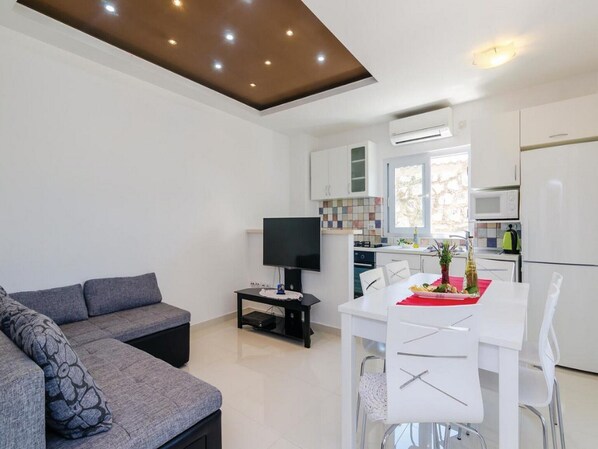 A modern living room with a grey sofa, a dining table with white chairs, a flat-screen TV, and a kitchen area with white cabinets and appliances.