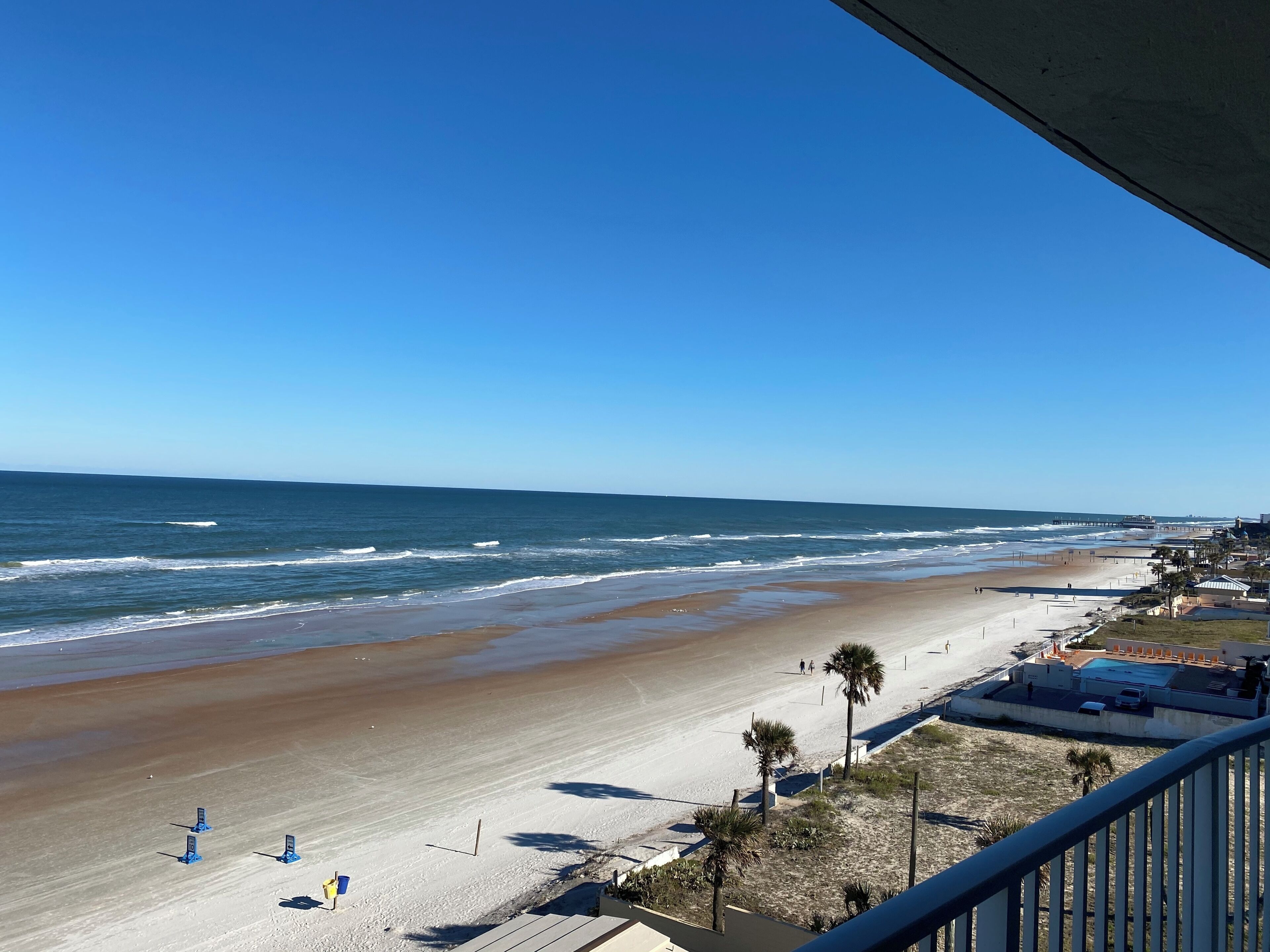 A beach with a pier in the distance, clear skies, and a few people walking on the sand.