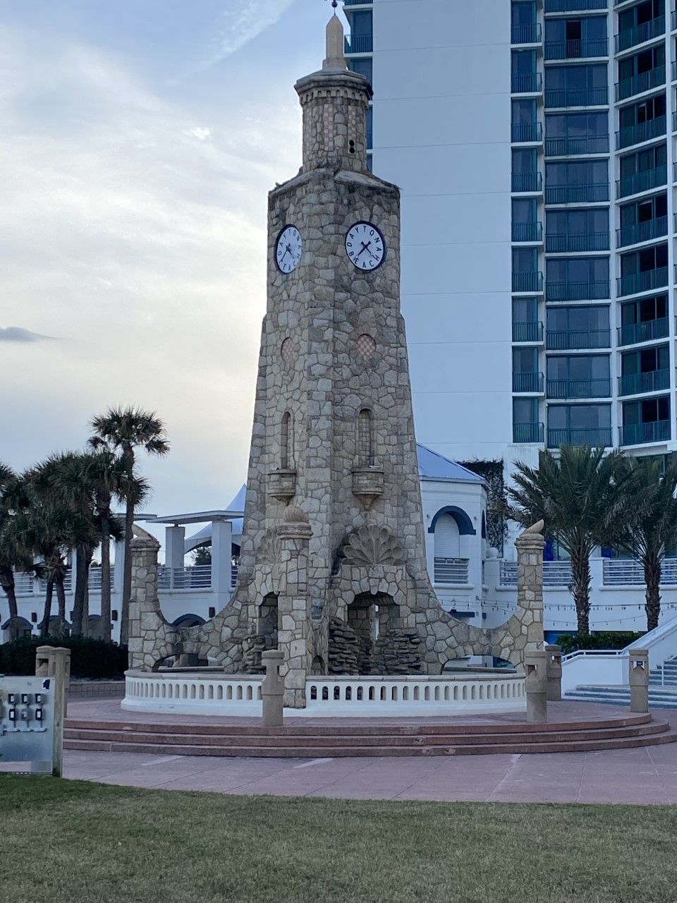 A stone clock tower with two clocks, surrounded by a white fence and palm trees.