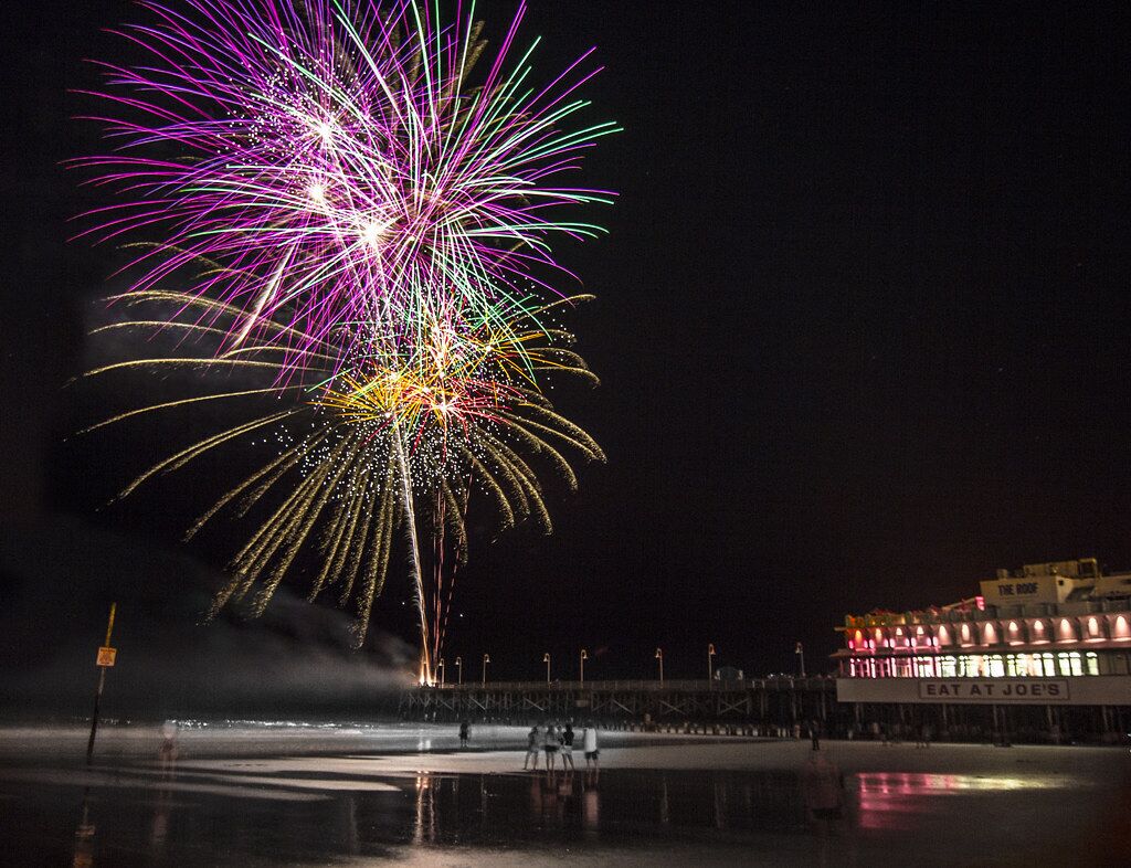 A nighttime scene with fireworks over a waterfront area, a pier, and a building with a sign reading "EAT AT JOE'S".