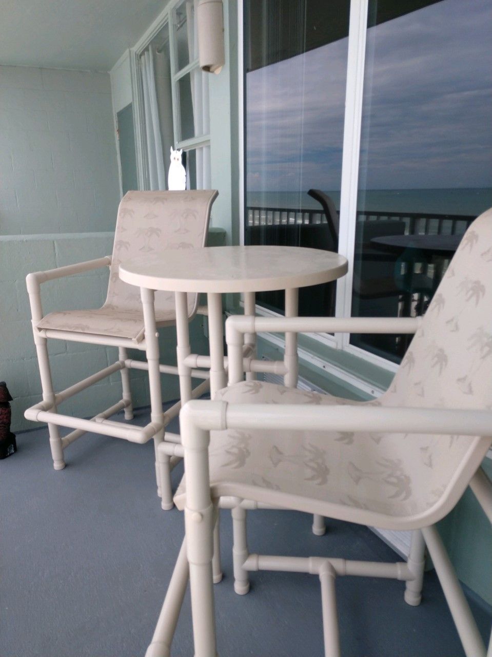 A balcony with two white chairs and a small round table, a window, and a view of the sea.