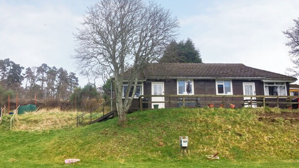 Ein zweigeschossiges Holzhaus mit geneigtem Dach, einer Holzdiele und einem großen Baum davor.