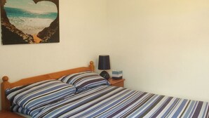Ein Bett mit gestreifter Bettwäsche, ein Nachttisch mit Lampe, eine Kiste und ein Wandbild mit Herz und Strandmotiv.