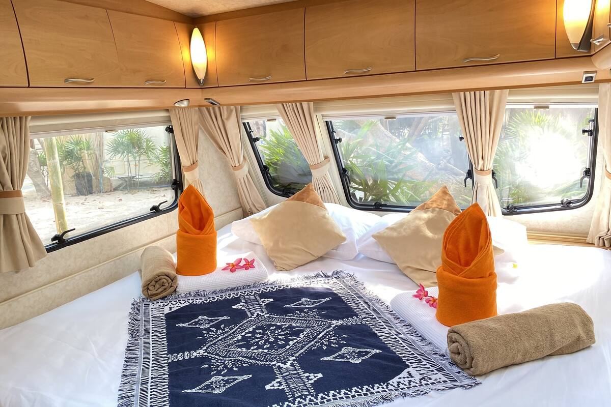 A cozy bed with a patterned throw, neatly folded towels, and a view of greenery through the windows.