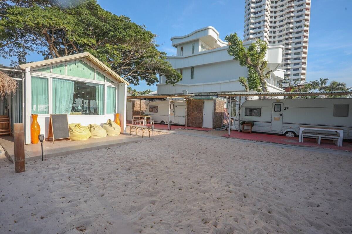A beachside RV park with a sandy area, a small building with a window, and a tall residential building in the background.