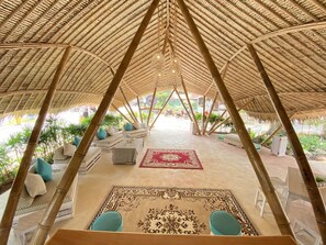 An open-air lounge with a thatched roof, cushioned seating, and a patterned rug. - Beach House Camp (Sattahip)