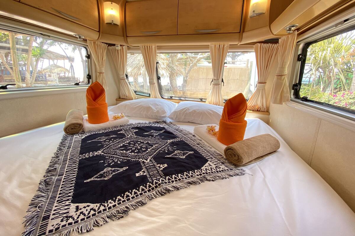 A bed with a patterned blanket, pillows, and towels, with curtains and a window view.