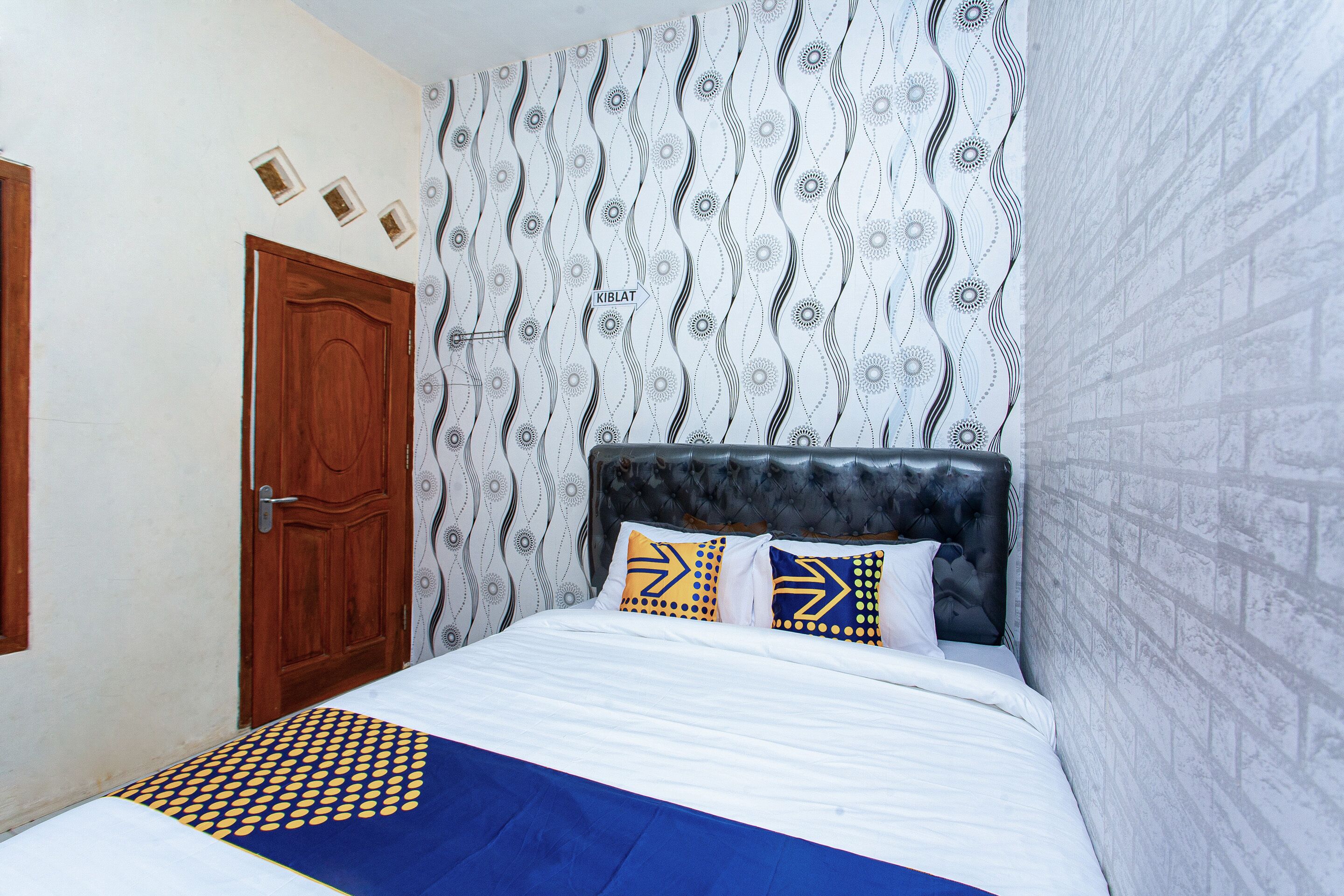 Un dormitorio con papel tapiz estampado, una puerta de madera, un cabecero acolchado y una cama con almohadas estampadas.