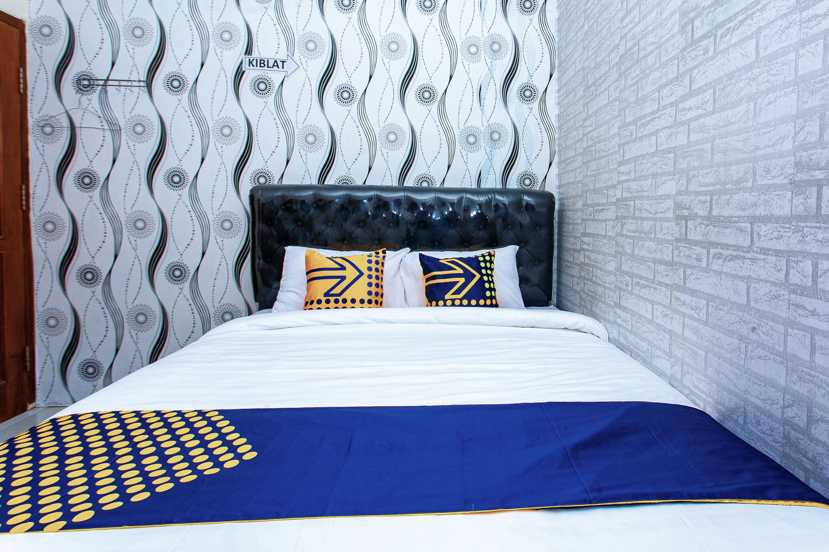 Een net opgemaakt bed met een blauw-gouden deken en gestreepte kussens, tegen een muur met golvend behang en een baksteenstructuur.