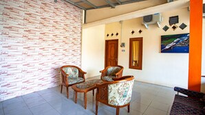 A room with a brick-patterned wall, two wooden chairs with floral upholstery, a wooden table, and a painting on the wall. - SPOT ON 2875 Kost Pandawa Syariah (Cilacap)