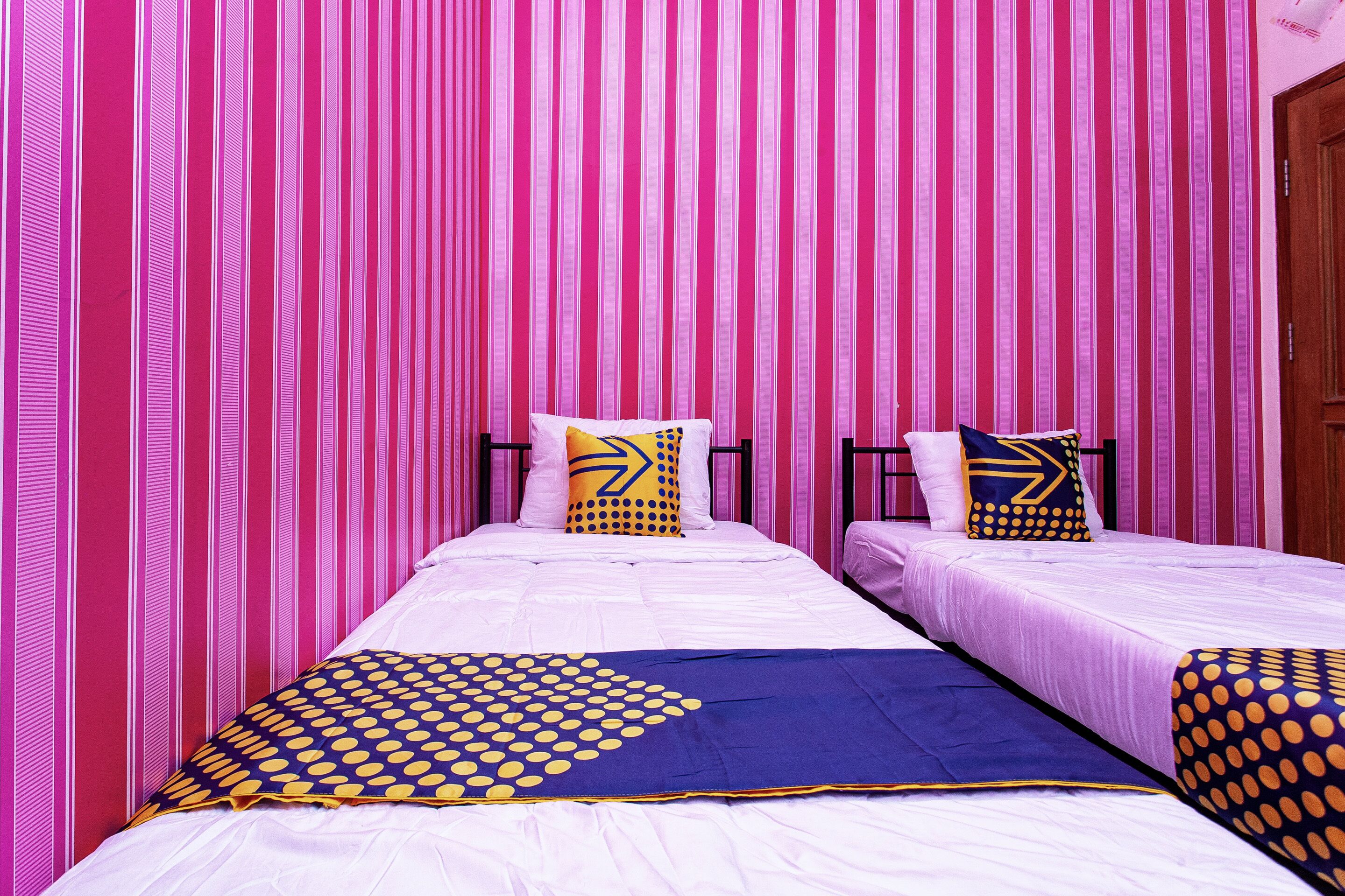 A room with two beds, each with a white mattress and a blue and yellow polka dot blanket. The walls are pink with vertical stripes.