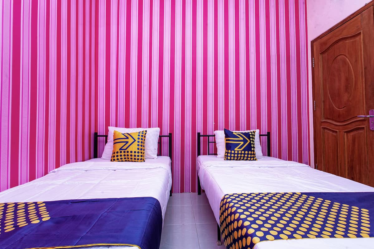 Een kamer met twee bedden, elk voorzien van een wit matras en een bedspreide met blauw en geel dessin. De muren zijn gestreept in roze en wit, en aan de rechterkant is een houten deur.