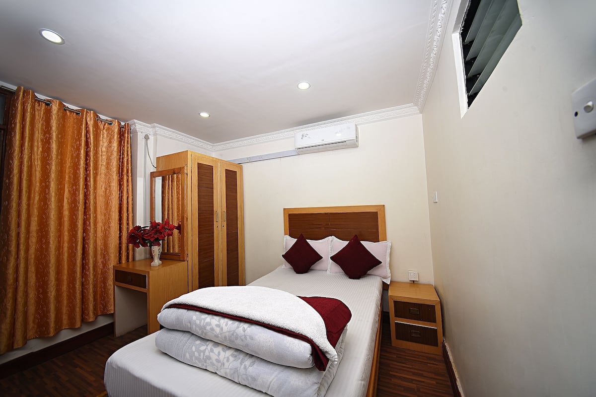 A hotel room with a bed, wooden headboard, bedside tables, a nightstand, a mirror, a window with curtains, an air conditioning unit, and a wall-mounted light fixture.