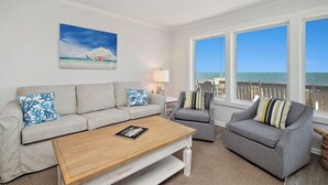 A living room with a sofa, armchair, and coffee table, a painting of sailboats, and a view of the ocean through large windows.