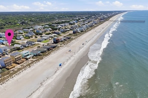 A coastal residential area with houses along the beach, a pier extending into the water, and a clear sky.