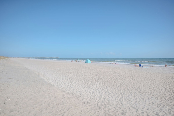 Une vaste plage de sable fin, un ciel dégagé, une jetée lointaine, ainsi que quelques personnes et des tentes.