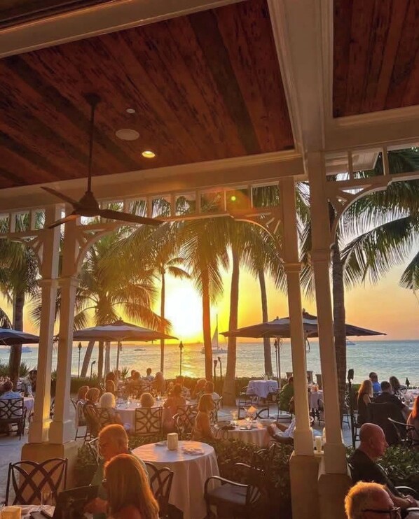 An outdoor dining area with tables and chairs, palm trees, and a view of the sunset over the ocean.