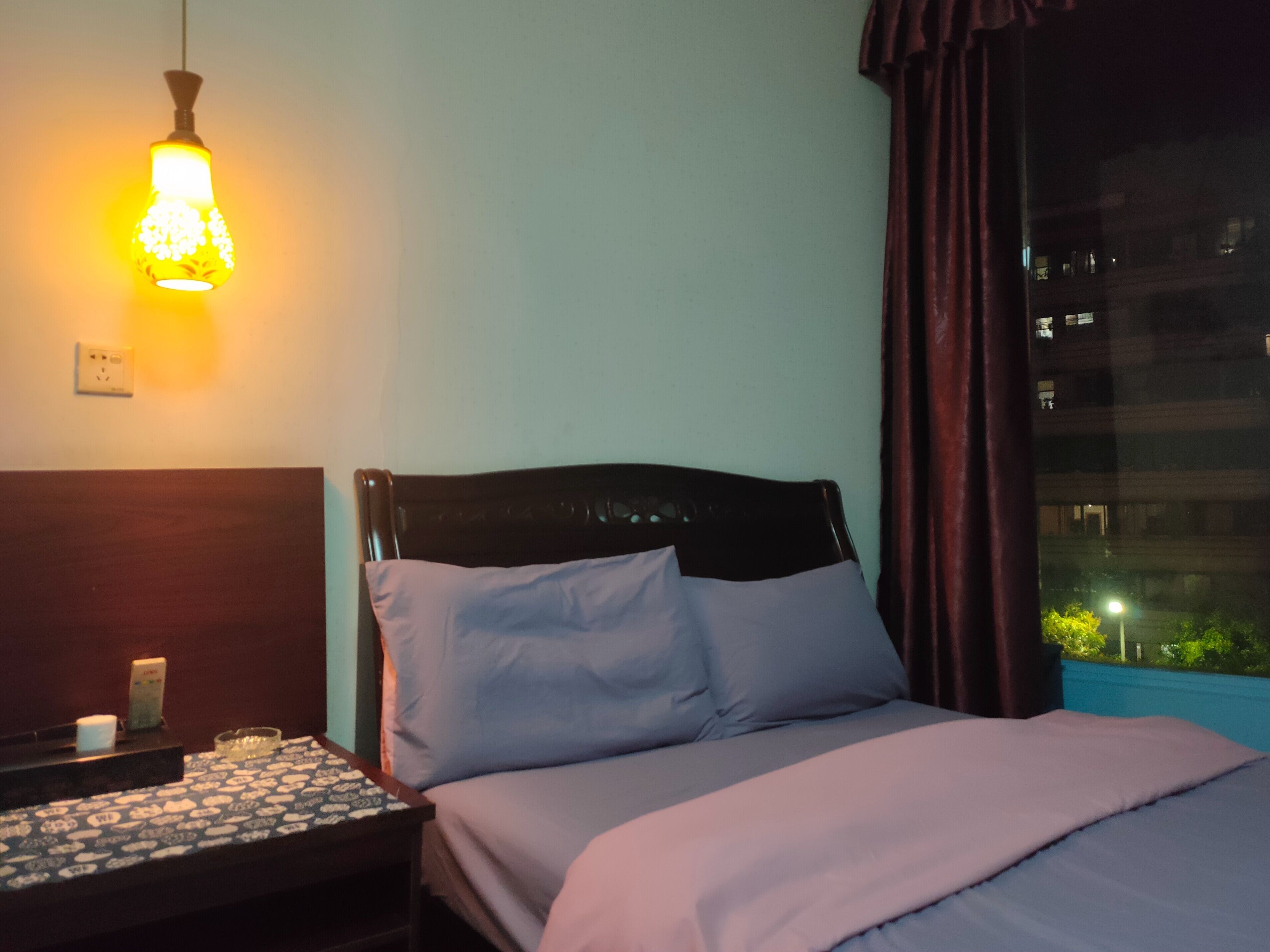 A hotel room with a bed, a nightstand, a lamp, and a window with a view of buildings.