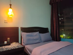A hotel room with a bed, a nightstand, a lamp, and a window with a view of buildings.