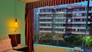 A hotel room with a bed, a nightstand, a lamp, a window with curtains, and a view of a multi-story building. - Foshan school age Youth Hostel (Foshan)
