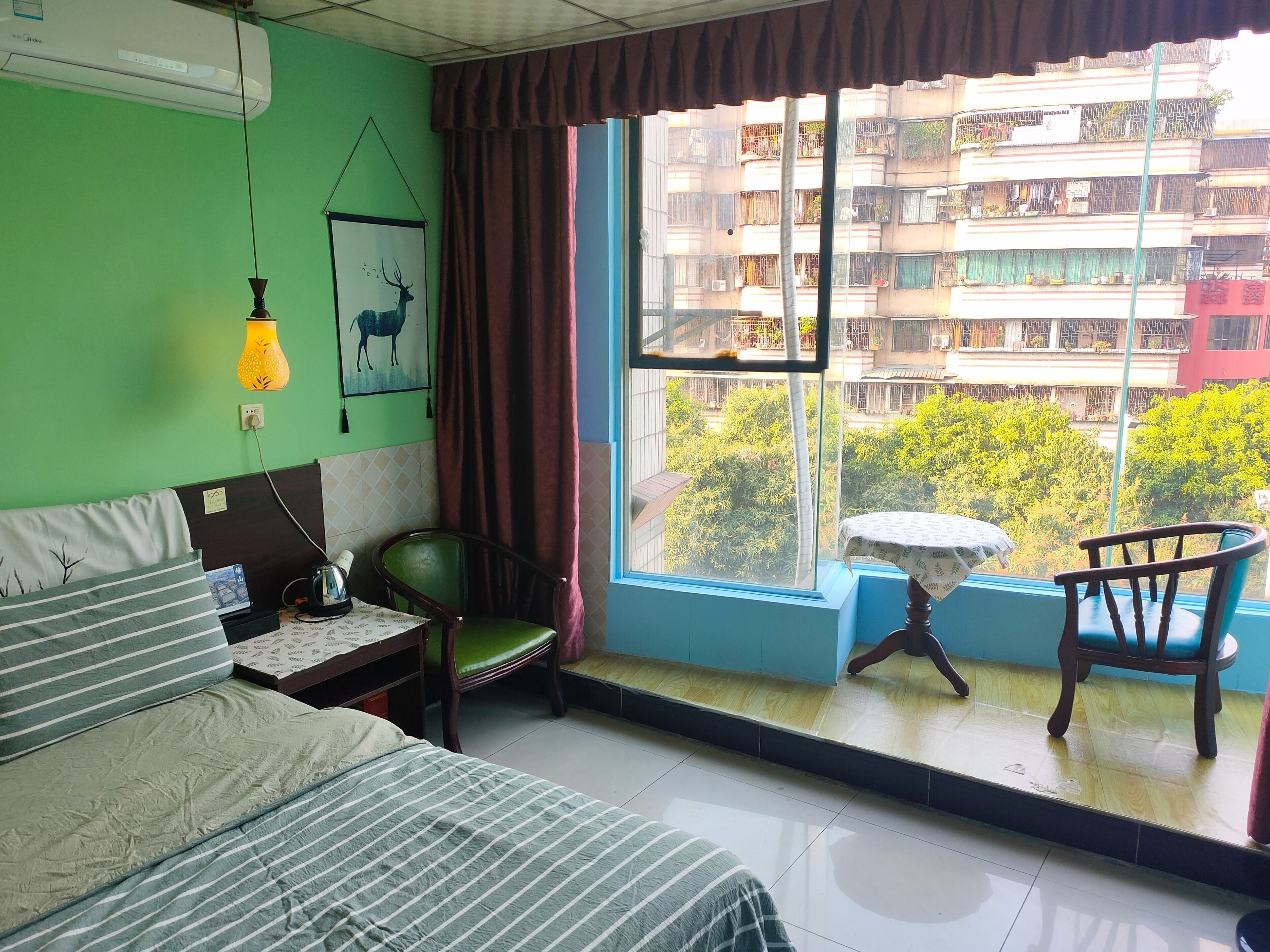 A bedroom with a bed, a chair, a small table, a window with a view of buildings and greenery, and a wall-mounted air conditioner.