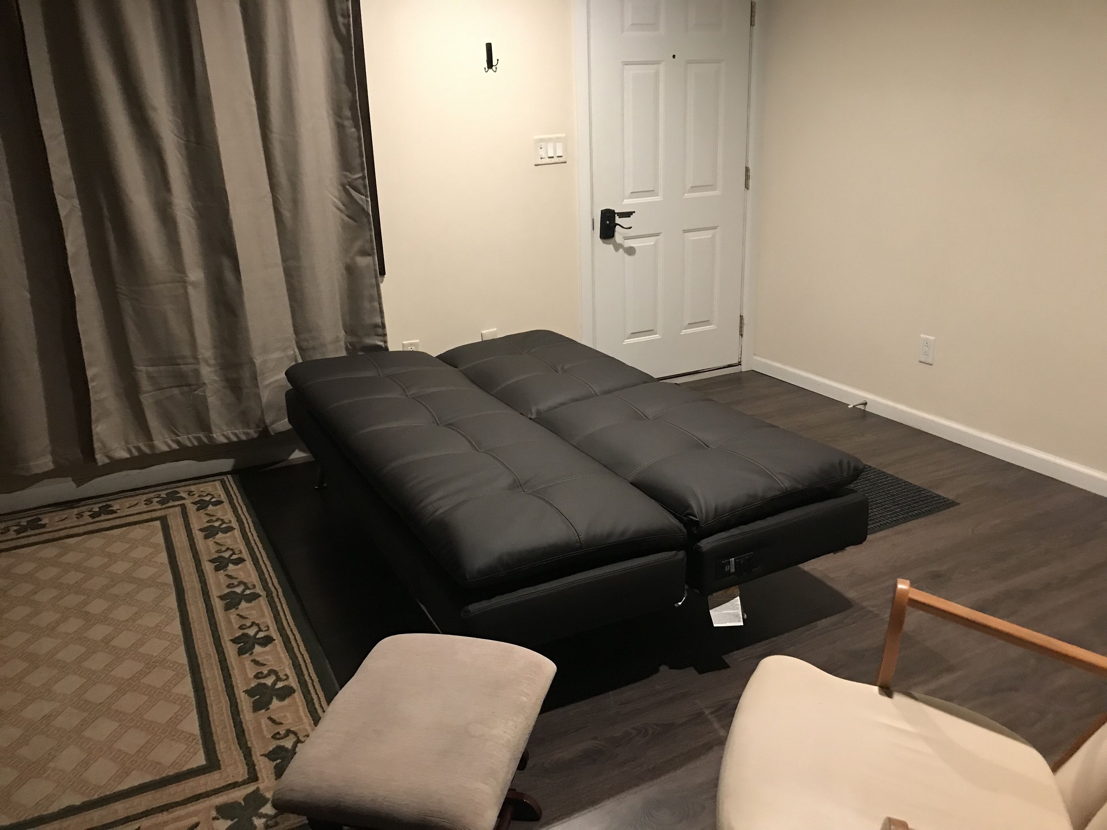 A room with a black sofa bed, a small beige chair, a patterned rug, and a white door.