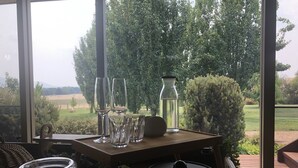 Uma mesa de madeira com copos transparentes, um jarro e uma panela em uma varanda com vista para árvores e campos.