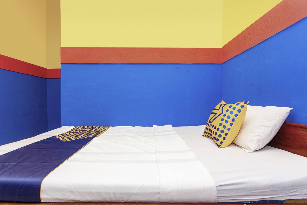 Uma cama com colcha branca e azul, um travesseiro amarelo com bolinhas azuis, e uma cabeceira de madeira.