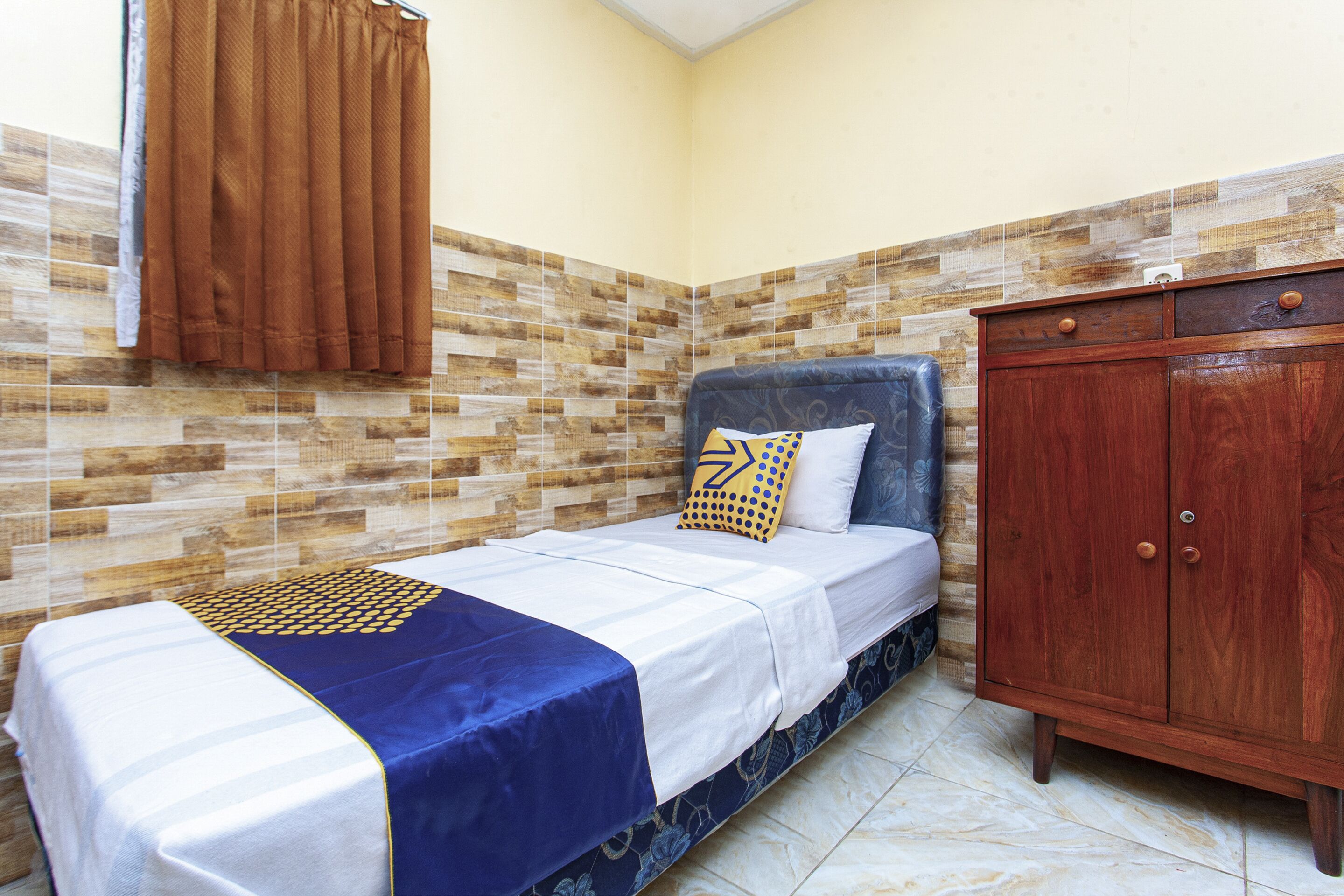 Une petite pièce, bien rangée, avec un lit, une armoire en bois et un mur orné d’un motif.