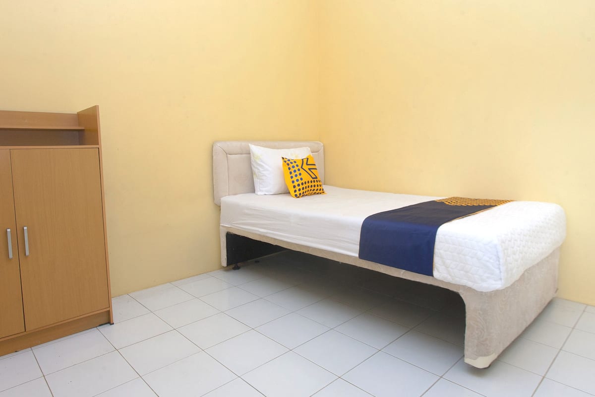 A single bed with a white mattress, a blue blanket, and a yellow pillow with a geometric pattern. There is a wooden wardrobe with two doors and a handle on each. The room has a plain yellow wall and white tiled flooring.